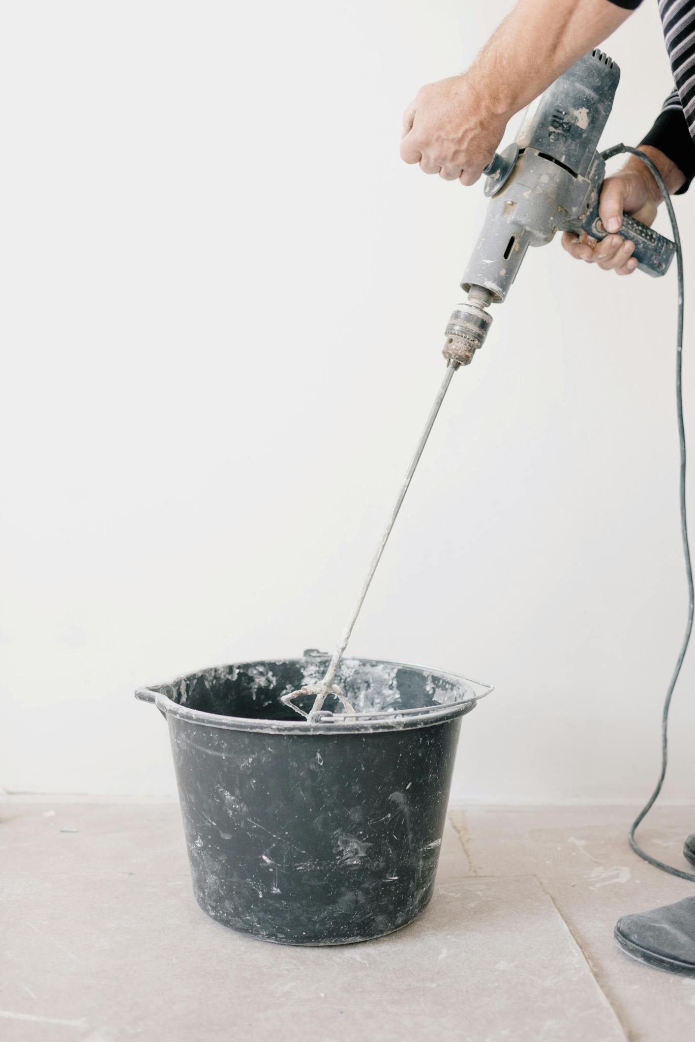 Worker using an electric drill to mix cement in a bucket during home renovation.